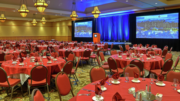 a large meeting room with red tables and chairs, a stage, and a large screen