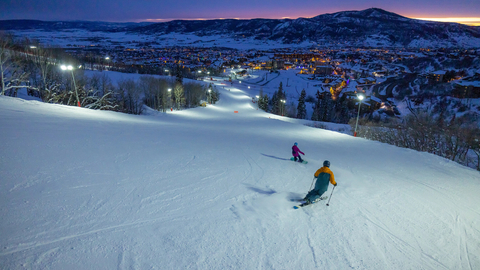 Two skiers skiing down a snowy slope at night with a town and mountains in the background.
