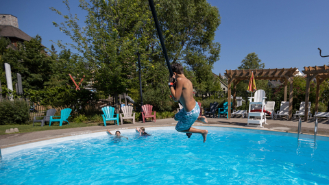 Child swinging into an outdoor pool at The Plunge! at Blue Mountain on a sunny day, surrounded by Adirondack chairs and families enjoying summer activities at the water park.