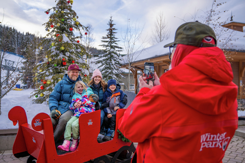 A family sits in Santa's sleigh in front of a decorated tree with ski slopes in the background.