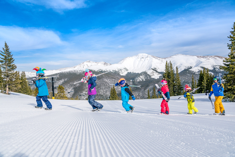 A group of four children and one adult walk across the snow while carrying skis on their shoulders at Winter Park Ski Resort in Colorado.