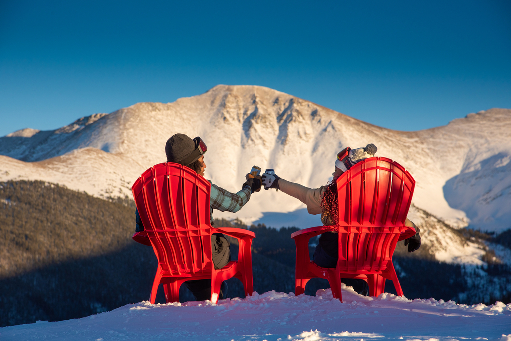 two people cheers with cans of beer while sitting in red adirondack chairs overlooking Parry Peak at Winter Park Resort